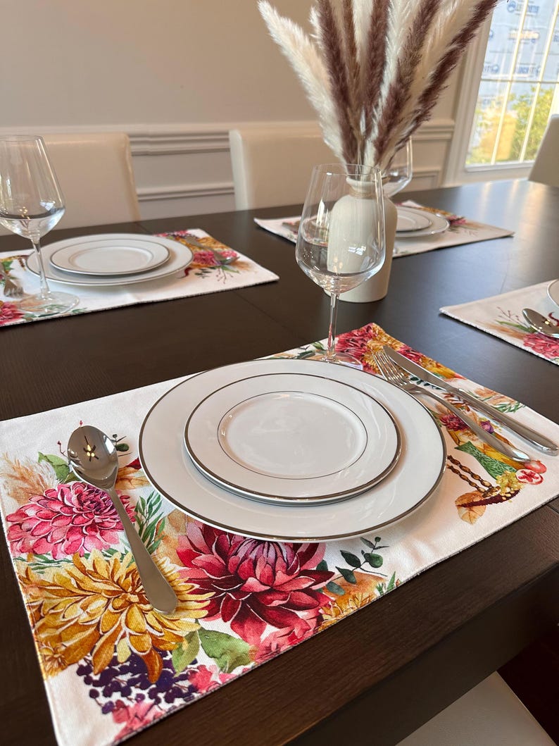 K&ouml;nnte beinhalten: Ein wei&szlig;er Platzteller mit einem Blumendesign mit rosa, gelben und violetten Blumen. Der Platzteller ist mit einem wei&szlig;en Teller, einer silbernen Gabel und einem Messer sowie einem silbernen L&ouml;ffel gedeckt.