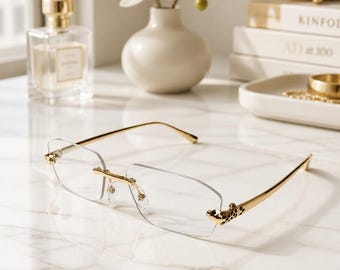 May include: Rimless eyeglasses featuring thin gold-tone metal temples and a bridge. The hinges are adorned with a decorative gold-tone leopard head motif with black enamel spots. The lenses are clear, rectangular, and polished at the edges.