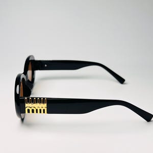 May include: Black sunglasses with round frames and gold accents on the temples. The lenses are a light brown color. The sunglasses are set against a white background.