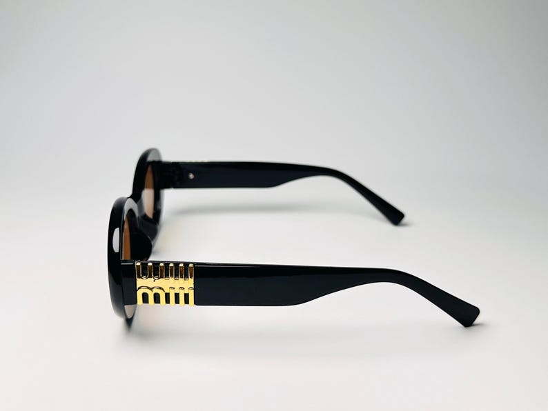 May include: Black sunglasses with round frames and gold accents on the temples. The lenses are a light brown color. The sunglasses are set against a white background.