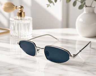May include: Oval-shaped sunglasses featuring a thin, polished silver metal frame and dark, solid-colored lenses. The eyewear has slender temples and a bridge connecting the two oval rims, providing a sleek and streamlined appearance for the wearer.