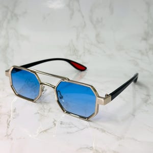 May include: Stylish sunglasses with a gold-colored frame and blue lenses. The glasses have a unique octagonal shape and black arms with red accents on the tips. The sunglasses are designed for a modern look.