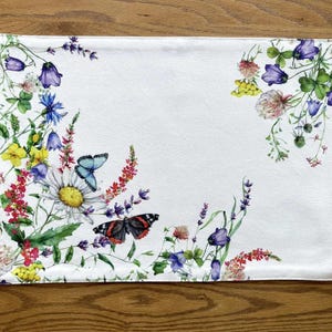 K&ouml;nnte beinhalten: Wei&szlig;e Tischsets aus Leinen mit einem Blumendesign mit Schmetterlingen, Wildblumen und gr&uuml;nen Bl&auml;ttern. Die Blumen sind in den Farben Rosa, Lila, Gelb und Blau gehalten.