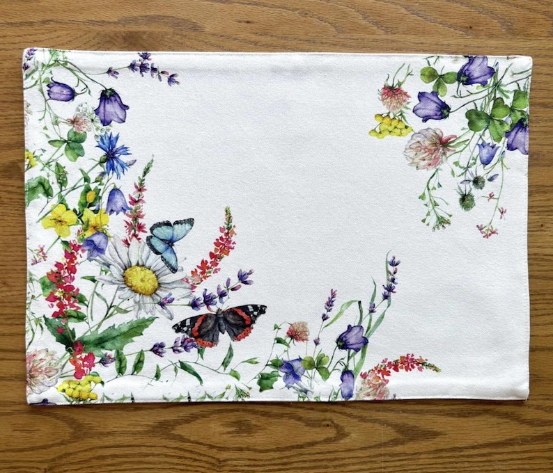 K&ouml;nnte beinhalten: Wei&szlig;e Tischsets aus Leinen mit einem Blumendesign mit Schmetterlingen, Wildblumen und gr&uuml;nen Bl&auml;ttern. Die Blumen sind in den Farben Rosa, Lila, Gelb und Blau gehalten.