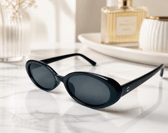 May include: A pair of black oval-shaped sunglasses with dark lenses and a glossy finish. The frames feature a simple, rounded design with thin temples and small silver-toned metal accents at the hinges, providing a clean and uniform appearance.