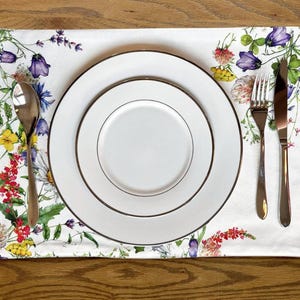 K&ouml;nnte beinhalten: Ein wei&szlig;er Blumen-Platzteller mit einem Satz von drei wei&szlig;en Tellern und silbernem Besteck. Der Platzteller zeigt ein Aquarell-Design mit verschiedenen Blumen in den Farben Rosa, Gelb, Blau und Gr&uuml;n.