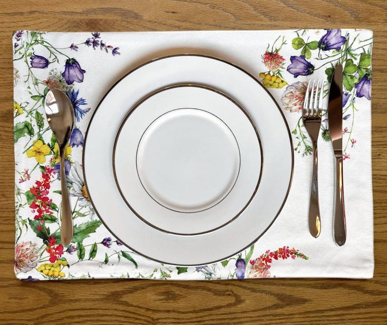 K&ouml;nnte beinhalten: Ein wei&szlig;er Blumen-Platzteller mit einem Satz von drei wei&szlig;en Tellern und silbernem Besteck. Der Platzteller zeigt ein Aquarell-Design mit verschiedenen Blumen in den Farben Rosa, Gelb, Blau und Gr&uuml;n.