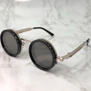 May include: Round sunglasses with black rims and dark grey lenses. The frames are silver-toned with black temples. The lenses have a dial with the text "MIN" and "MAX". The sunglasses are on a white marble surface.