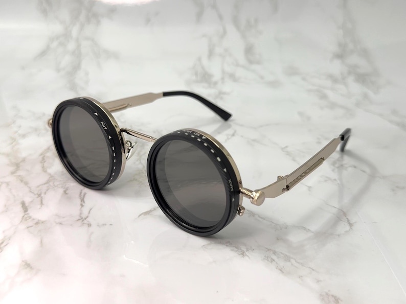 May include: Round sunglasses with black rims and dark grey lenses. The frames are silver-toned with black temples. The lenses have a dial with the text "MIN" and "MAX". The sunglasses are on a white marble surface.