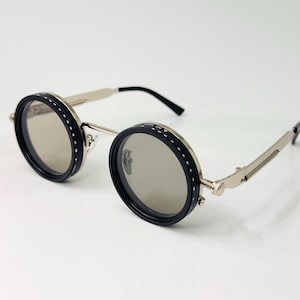 May include: Round sunglasses with black frames and silver arms. The lenses are a light grey colour. The frames have a stitched design around the lenses. The arms have a hinge design.
