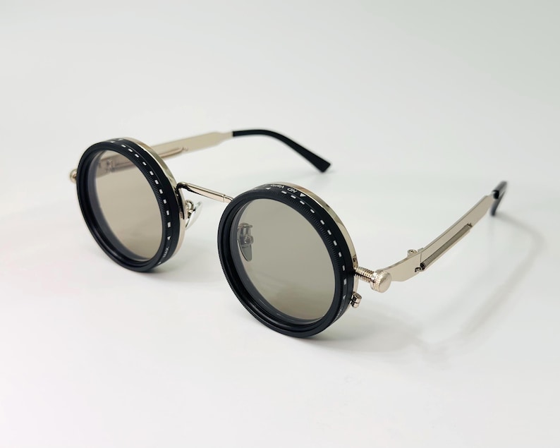 May include: Round sunglasses with black frames and silver arms. The lenses are a light grey colour. The frames have a stitched design around the lenses. The arms have a hinge design.