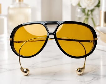 May include: Oversized aviator sunglasses featuring a thick black frame with a distinctive triangular bridge cutout and vibrant yellow-tinted lenses. The thin gold-toned metal temples curve downward, ending in smooth, rounded gold-toned ball tips.
