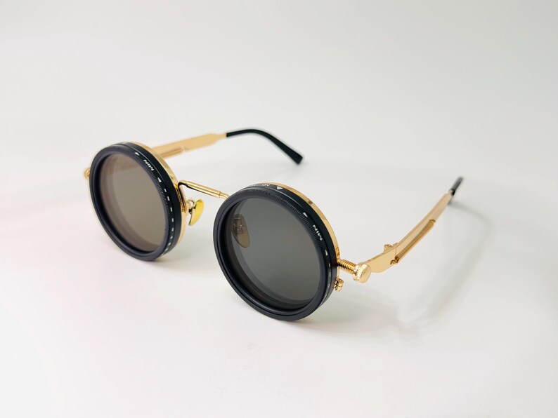 May include: Round sunglasses with black frames, gold-coloured temples, and dark lenses. The frames have a black outer ring with white stitching. The temples are gold with black earpieces. The sunglasses are set against a white background.