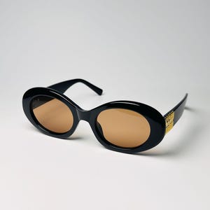 May include: Black oval sunglasses with brown lenses. The sunglasses have a thick frame and gold-colored detailing on the temples. The lenses are a warm brown color, and the frame is glossy black.