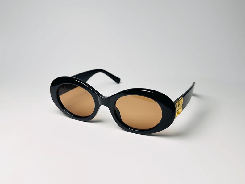 May include: Black oval sunglasses with brown lenses. The sunglasses have a thick frame and gold-colored detailing on the temples. The lenses are a warm brown color, and the frame is glossy black.