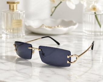 May include: Rimless rectangular sunglasses featuring dark gray tinted lenses and thin gold-tone metal temples. The bridge and hinges are constructed from polished gold-tone metal, connecting the lenses with small screws at the outer edges.