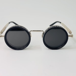 May include: Circular sunglasses with black frames and dark lenses. The frames feature a silver bridge and temples. The lenses are set in black casings. The design has a steampunk style.
