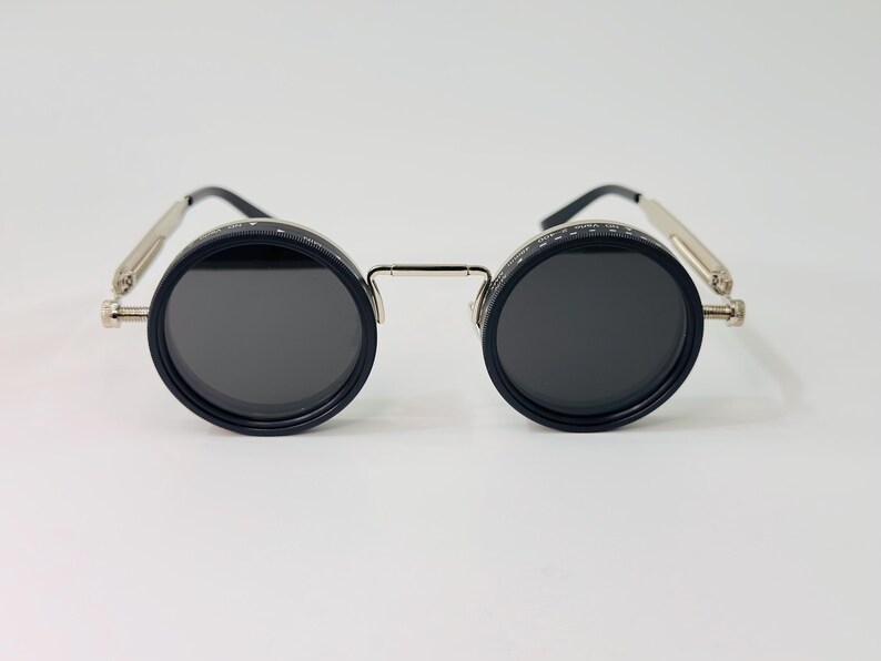 May include: Circular sunglasses with black frames and dark lenses. The frames feature a silver bridge and temples. The lenses are set in black casings. The design has a steampunk style.