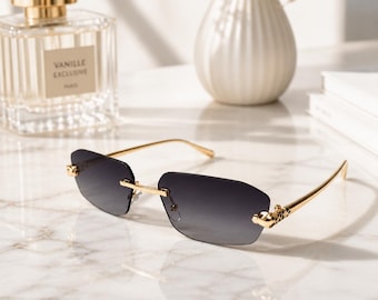 May include: Rimless sunglasses featuring rectangular gradient gray lenses and polished gold-tone metal temples. The hinges are detailed with a small, sculpted gold panther head design, providing a distinct metallic accent to the frame structure.