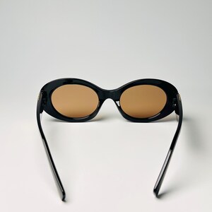 May include: Black oval sunglasses with brown lenses. The sunglasses have a thick frame and thin arms. The arms are extended, and the sunglasses are set against a white background. The lenses are a light brown color.