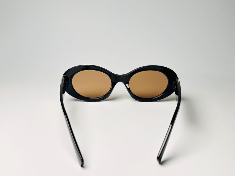 May include: Black oval sunglasses with brown lenses. The sunglasses have a thick frame and thin arms. The arms are extended, and the sunglasses are set against a white background. The lenses are a light brown color.