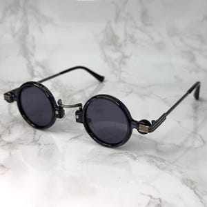 Puede incluir: Gafas de sol redondas con montura negra y lentes oscuras. Las gafas tienen un diseño de bisagra único y patillas negras delgadas. Las lentes son circulares y el estilo es vintage o retro.