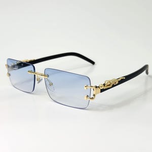 May include: Rimless sunglasses with light blue gradient lenses. The frames are gold-colored with black temples. A gold-colored jaguar design is on the temples. The sunglasses are rectangular in shape.