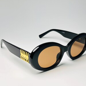 May include: Black oval sunglasses with gold accents on the temples and brown tinted lenses. The sunglasses have a thick frame and a classic, stylish design.