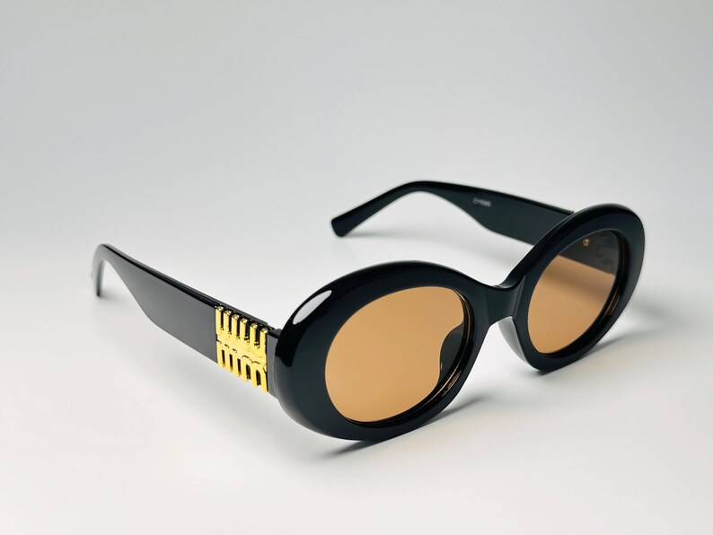 May include: Black oval sunglasses with gold accents on the temples and brown tinted lenses. The sunglasses have a thick frame and a classic, stylish design.