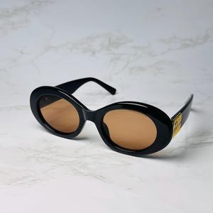 May include: Black oval sunglasses with brown lenses. The sunglasses have a thick frame and gold accents on the temples. The sunglasses are set against a white marble background.