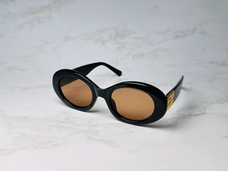 May include: Black oval sunglasses with brown lenses. The sunglasses have a thick frame and gold accents on the temples. The sunglasses are set against a white marble background.