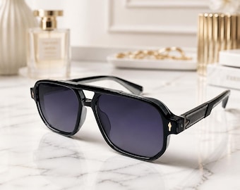 May include: Black sunglasses featuring a thick frame with a double bridge design and dark tinted lenses. The temples have a textured finish, and small gold-toned diamond-shaped accents are visible on the front corners of the frame.