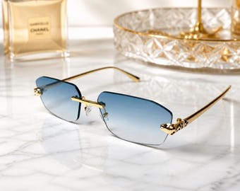 May include: Rimless sunglasses featuring gradient blue lenses and gold-tone metal temples. The hinges are adorned with a small, detailed gold leopard head motif with black spots, connecting the frame to the slender, polished gold arms.