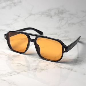 May include: Black aviator sunglasses with orange-tinted lenses. The sunglasses have a double bridge and a thick frame. The temples feature a small arrow design. The sunglasses are set against a white marble background.