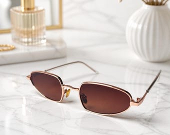 May include: Oval-shaped sunglasses featuring a metallic rose gold frame and dark brown gradient lenses. The eyewear has thin, slender temples and adjustable nose pads, showcasing a sleek, lightweight construction with a polished finish.