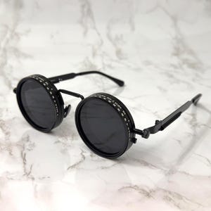 以下が含まれることがあります： スチームパンクデザインの黒い丸いサングラス。サングラスは黒い金属フレームと濃い色のレンズです。レンズの縁には装飾的な模様があります。サングラスのアームは細くて黒いです。