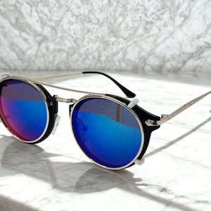 May include: Round sunglasses with a black frame and silver accents. The lenses have a vibrant blue and red gradient. The sunglasses feature a double bridge and a sleek, modern design, perfect for sunny days.