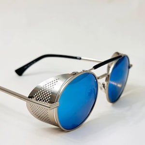 May include: Steampunk-style sunglasses with round, blue-tinted lenses and silver frames. The glasses feature side shields with a perforated design and black temples. The overall aesthetic is a blend of vintage and futuristic elements.