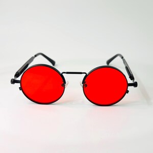 May include: Round sunglasses featuring vibrant red lenses and a thin black metal frame. The eyewear includes a straight bridge connecting the circular lenses and slender black temples with curved ends, providing a distinct and symmetrical appearance.