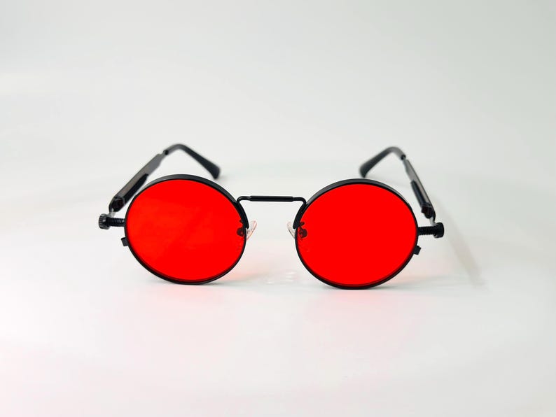 May include: Round sunglasses featuring vibrant red lenses and a thin black metal frame. The eyewear includes a straight bridge connecting the circular lenses and slender black temples with curved ends, providing a distinct and symmetrical appearance.
