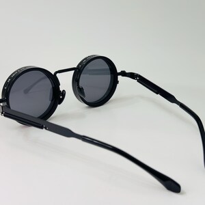 May include: Black round sunglasses with dark grey lenses. The sunglasses have a black metal frame and arms. The lenses are circular and the frame has a unique design. The arms are thin and curve at the ends.