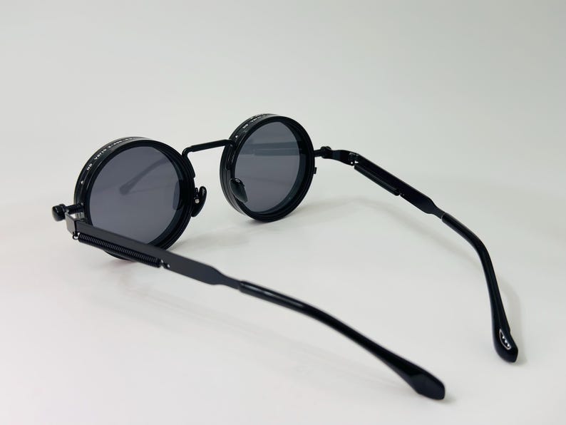 May include: Black round sunglasses with dark grey lenses. The sunglasses have a black metal frame and arms. The lenses are circular and the frame has a unique design. The arms are thin and curve at the ends.