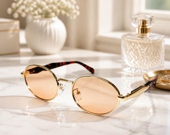 May include: Oval sunglasses featuring a thin gold-tone metal frame and light peach-tinted lenses. The temples are crafted with a brown and black tortoiseshell pattern, providing a contrast to the metallic front and the clear nose pads.