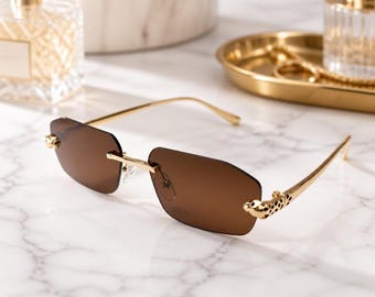 May include: Rimless sunglasses featuring rectangular brown lenses and gold-tone metal temples. The hinges are adorned with a detailed gold leopard head motif, and the bridge is a thin gold metal bar connecting the two lenses securely.