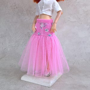 May include: A doll wearing a white crop top and a pink skirt. The skirt has a floral pattern on the waistband and a layered tulle design. The doll is standing on a white wooden block.