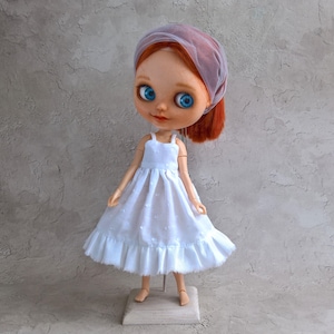 May include: A handmade doll with red hair and bright blue eyes, wearing a white dress with a ruffled hem. The doll has a sheer, lavender head covering. The doll is standing on a wooden base with a metal stand.