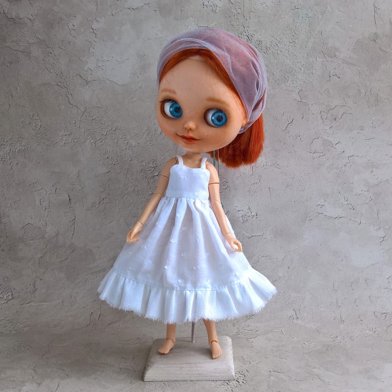 May include: A handmade doll with red hair and bright blue eyes, wearing a white dress with a ruffled hem. The doll has a sheer, lavender head covering. The doll is standing on a wooden base with a metal stand.