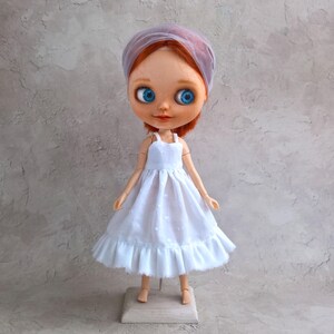 May include: A doll with red hair, blue eyes, and a white dress with a ruffled hem. The doll wears a sheer, light purple head covering. The doll is standing on a small wooden stand.