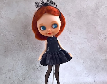 May include: A doll with red hair and blue eyes, wearing a black dress and tights. The doll has a black lace bow headband. The dress features a ruffled skirt. The doll stands on a white stand.