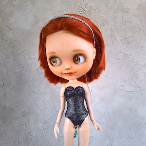 Könnte beinhalten: Eine Puppe mit rotem Haar, Sommersprossen und großen, ausdrucksstarken Augen. Die Puppe trägt einen schwarzen, glitzernden Badeanzug und ein silbernes Stirnband. Die Merkmale der Puppe sind detailliert, und die Gesamtästhetik ist skurril und künstlerisch.
